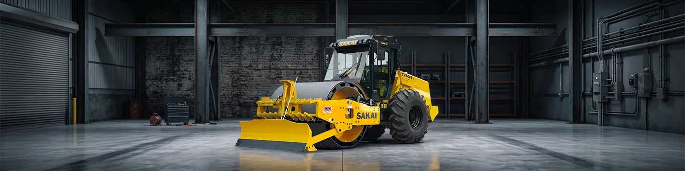 A new SAKAI SV544-1 84 inch soil compactor with strike off blade, cab, and smooth shell kit is dimly lit inside an industrial facility.