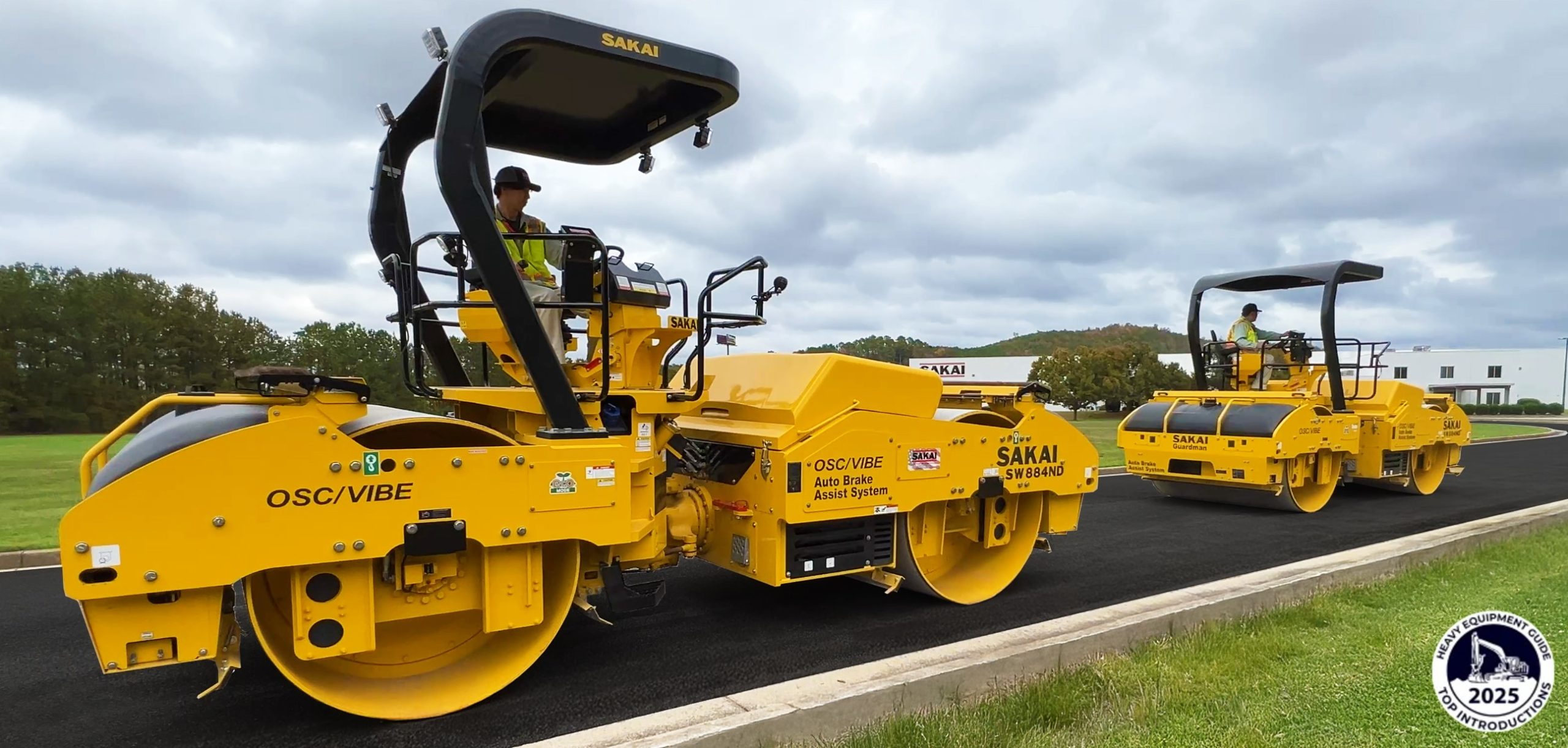 Two SAKAI SW884ND asphalt rollers equipped with the new Guardman Echelon Mode roll in tandem formation in front of the SAKAI factory. A Heavy Equipment Guide 2025 Top Introductions logo is in the bottom right of the image.