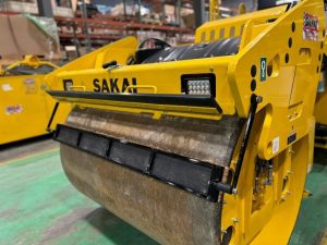 Coco mat assembly mounted on a SAKAI SW774 66 inch 11 ton asphalt roller with mat in down position and scraper in up position.