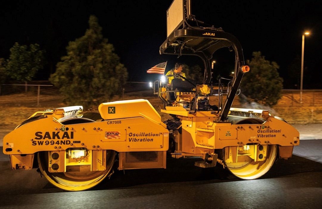 A Sakai SW994ND 79 inch highway class double drum oscillatory asphalt roller on paving job at night with LED drum lights.